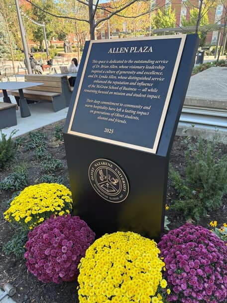 An image of an outdoor sign surrounded by purple and yellow flowers.